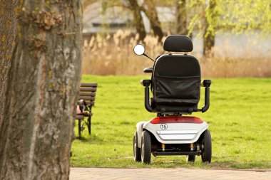 Bir yaz parkında ağacın yanında elektrikli tekerlekli sandalye duruyor. Kendi ayakları hareket etmekte zorlanan engelli ve yaşlılara yapılan yardımların otomasyonu ve mekanizması. İhtiyaç sahiplerine bakıyorum.