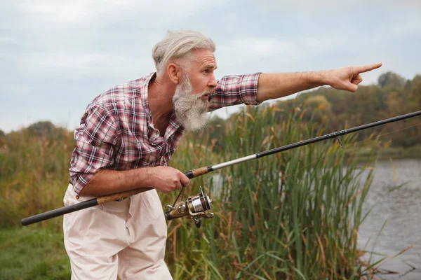 Pescador Sênior Apontando Para Longe Enquanto Pesca Com Sua Haste — Fotografia de Stock