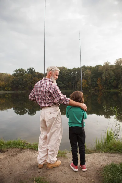 湖で一緒に釣りながら 彼の孫を抱きしめる上級漁師の垂直フル長さのリアビューショット — ストック写真