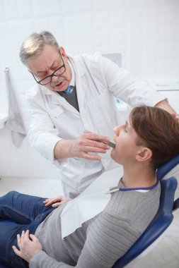 Kliniğinde çalışan yaşlı bir dişçinin dikey çekimi hastanın dişlerini kontrol ediyor.