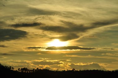 Yoğun bir orman bölgesinin ardında batan bir güneşin resmi. Ardından dağlar..