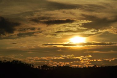 Yoğun bir orman bölgesinin ardında batan bir güneşin resmi. Ardından dağlar..