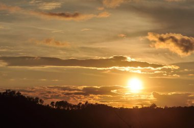 Yoğun bir orman bölgesinin ardında batan bir güneşin resmi. Ardından dağlar..