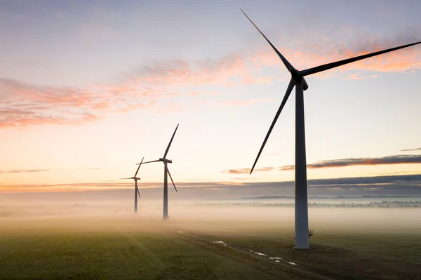 Sabahın erken saatlerinde İngiliz kırsalında gün doğumunda üç rüzgar türbininin hava görüntüsü.