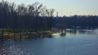 Sunny day view of the Dnieper river flowing through Kyiv with the Motherland Monument and the Ukrainian flag visible on the horizon, silhouetted against a clear sky during early spring