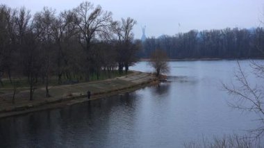 One solitary fisherman is seen on the concrete riverside of the Dnipro river in Kyiv, surrounded by serene water mirroring the leafless trees and the iconic Motherland Monument in the far distance