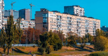 Large residential building standing prominently above a lush green park, where people are walking and cycling, with modern high rises and construction cranes in the background under a clear sky