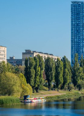 Urban river park contrasting modern high rise and residential buildings with lush green trees, a popular recreation area offering pedal boat rentals for relaxing summer activities