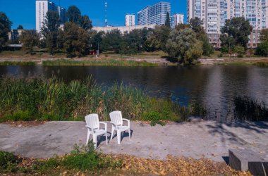 White plastic chairs on a concrete riverbank, urban park providing a quiet escape with a river and city buildings, contrasting nature and architecture, depicting relaxation and autumn