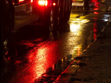 Wet asphalt street reflecting the bright red taillights of a truck and yellow headlights of an approaching car, creating dynamic light patterns on the dark, rain soaked road surface
