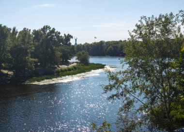 Mother Ukraine monument dominates the Kyiv skyline, overlooking the Dnieper with sunlit reflections, autumn trees along the banks and a distant Ukrainian flag fluttering gently