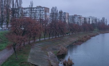 Nehir seti, sonbahar yapraklı bir sıra ağaç, beton bir istinat duvarı ve bulutlu bir gökyüzünün altında uzak apartman blokları olan bir yaya yolunu gösteriyor.