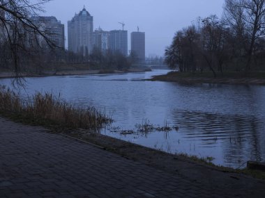 Şehir parkı nehir manzarası soğuk, bulutlu bir günde, çıplak ağaçlar, uzaktaki yüksek binalar, ve koyu asfalt bir set, kasvetli bir sinema havası yaratır.