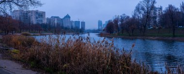 Şehir nehri, bulutlu gökyüzü altındaki modern ve eski şehir binalarının önünden akıyor. Ön planda kuru sazlıklar kasvetli ve sakin bir atmosferi yansıtıyor.