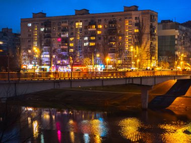 Şehir köprüsü, geceleri yüksek apartman binalarının altındaki bir nehri kaplıyor, karanlık suyu yansıtan pencere ve işyerlerinin parlak ışıkları, renkli bir şehir akşam manzarası yaratıyor.