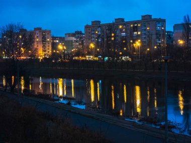 Alacakaranlıktaki şehir manzarasında aydınlık pencereleri, sokak lambaları ve sakin nehir yüzeyinde parlak sarı ve mavi yansımaları olan konut binaları yer alıyor.