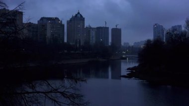 Kasvetli bir alacakaranlık sırasında Kyiv 'in modern binalarının Dnipro Nehri' nin sakin sularındaki yansımasını gösteren dramatik ve karamsar şehir manzarası gizemli ve atmosferik bir manzara yaratıyor.