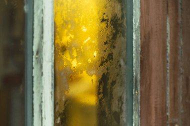 Donmuş, desenli camlı ve yıpranmış ahşap çerçeveli aşınmış pencere camının soyut görüntüsü, karamsar, klasik atmosferik bir sahne için sıcak altın ışık ve tozu yansıtıyor.