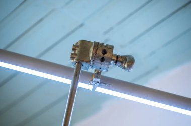 Endüstriyel metal kelepçe, parlak floresan lamba tüpünün yanında, mavi tavan arka planı, eklenti, güvenlik ve endüstriyel çözümleri temsil eden bir boruyu güvenli bir şekilde tutuyor.