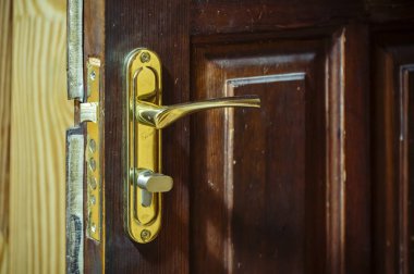 Zarar görmüş ahşap kapı, yerinden oynanmış altın bir kol ve görünür kilit mekanizması, güvenlik ihlali, haneye tecavüz veya tamir gereksinimleri gibi kavramlar içeriyor.