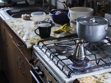 Sıcak bir ev mutfağı sahnesi, gaz ocağında kahve demleyen metal bir kafeterya, lavabo, çeşitli tencereler ve günlük yaşamı temsil eden desenli bir tezgah.