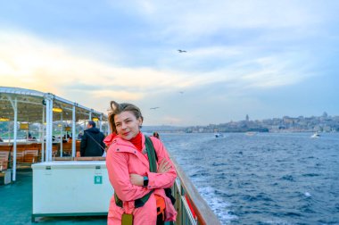 Boğaz feribotunda duran kadın, kollarını kavuşturup gülümsüyor ve denizin üzerinde uçan martılarla Galata Kulesi de dahil olmak üzere İstanbul şehir manzarasının tadını çıkarıyor.