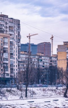 Şehir manzarası eski binaları, çıplak ağaçları ve modern yüksek binalarla çerçevelenmiş karla kaplı caddeleri ve bulutlu bir kış gökyüzünün altında çalışan vinçleri yakalıyor.