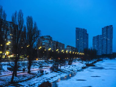 Kyiv 'in kışın alacakaranlık vakti kent manzarası konut binaları, karla kaplı bir nehir kıyısı, çıplak ağaçlar ve insanların yürüdüğü bir yolu aydınlatan sokak lambaları.