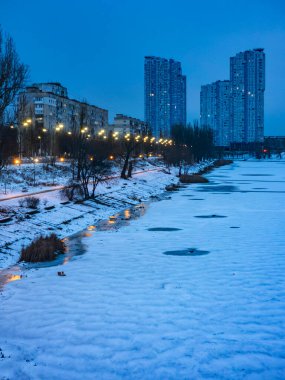 Buzlu bir nehrin, sokak lambalarıyla aydınlatılan karlı bir set boyunca kıvrılıp durduğu, modern yüksek binalar ve Sovyet döneminden kalma binaların koyu mavi gökyüzüne karşı durduğu kış şehirleri.