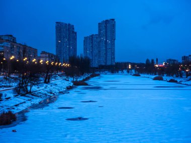 Şehir karla kaplı donmuş bir nehre ev sahipliği yapıyor. Şehir binaları ve nehir boyunca aydınlatılmış sokak lambaları sakin mavi bir alacakaranlık kentsel manzara sunuyor.