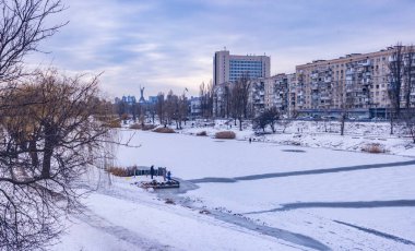 Kış panoraması donmuş bir kanalı, çıplak ağaçları, yerleşim binalarını ve bulutlu bir gökyüzünün altındaki Ukrayna Ana anıtını gösteren karla kaplı bir manzara.