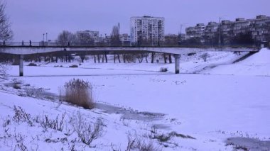 Donmuş bir nehrin üzerinde beton bir köprüde yürüyen yayalar ve arka planda bir şehir silüeti.
