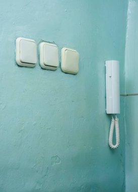 Üç ışık düğmesi ve eski beyaz telli bir dahili telefon yıpranmış, desenli su renkli bir duvara monte edilir ve yerleşim yerlerindeki günlük işlevsel elementleri gösterir.