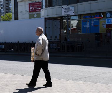 Kyiv, Ukrayna - 12 Mayıs 2022: Yaşlı bir adam kaldırımda yürüyor, bir çanta taşıyor, güçlü gölgeler ve hareketli bir şehir ortamında bina yansımaları taşıyor