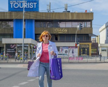 Kyiv, Ukrayna - 12 Mayıs 2022: Olgun bir kadın, elinde mor bir alışveriş torbası ve bir su şişesi ile şehrin arka planında binalar bulunan bir caddeden karşıya geçiyor.