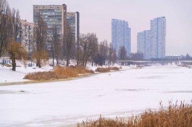 Donmuş nehir yüzeyi ve kuru sazlıklarla kaplı toprak seti soğuk kış havasını yansıtıyor, şehir binaları ve arka planda bir köprü bulunuyor.