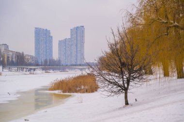 Kyiv 'de kış nehir manzarası kısmen donmuş bir kanal, karla kaplı kıyılar ve yerleşim yerlerinin yüksek binaları ve gri gökyüzünün altında yaya köprüsü ile çevrili çıplak söğüt ağaçları.