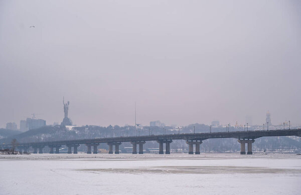 Kyiv cityscape showing the Paton Bridge spanning the Dnieper River, with a monumental statue and other city buildings visible on the rolling hills, all shrouded in a winter fog