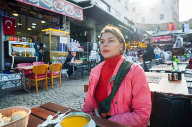 Bir kadın gezgin açık hava kafeteryasında oturmuş, yemeğin tadını çıkarıyor ve hareketli bir Türk pazar bölgesinin hareketli sokak yaşamını ve yerel atmosferini gözlemliyor.