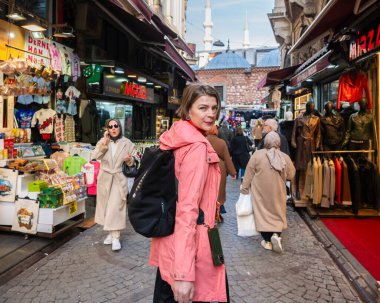 Sırt çantalı bir kadın İstanbul 'da hareketli dar bir caddede yürüyor, yerel dükkanları gözetliyor ve diğer insanlar bakınıyor, şehir pazarının canlı atmosferinin tadını çıkarıyor.