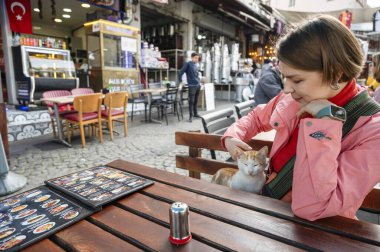 İstanbul 'da açık bir kafede oturan bir kadın, tek başına seyahat ederken dost canlısı bir zencefil sokak kedisini okşuyor, yerel sokak hayatının ve tarihi bir şehirde sakin bir anın tadını çıkarıyor.