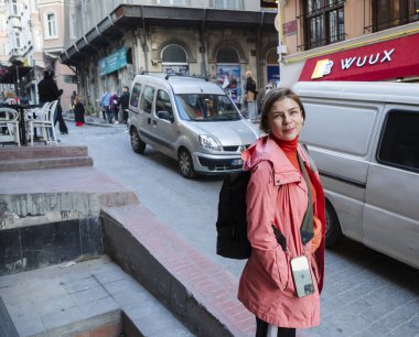 Yetişkin bir kadın tek başına seyahat ediyor, İstanbul 'da bir caddeyi keşfediyor, ceket giyiyor ve sırt çantası taşıyor, kameraya gülümsüyor, arka planda şehir binaları ve araçları var.