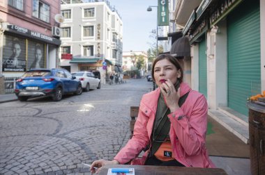 Pembe ceketli bir kadın, İstanbul 'da kaldırımlı bir caddede açık bir kafe masasında oturmuş, şehir keşfi sırasında kişisel bir mola veriyor.
