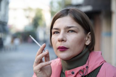 Şehir caddesinde sigara içen ve çevreyi izleyen bir kadın hareketli bir şehir ortamında düşünceli bir yalnızlık anı yaşıyor.