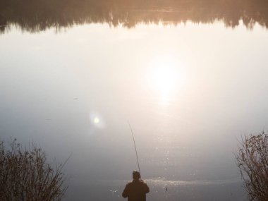 Kyiv, Ukrayna - 31 Mart 2023: Bir adam altın saatinde nehrin kenarında bir olta tutuyor, sıcak güneş ışığına karşı silueti var, sakin suya ve huzurlu yalnızlığa yansıyor.