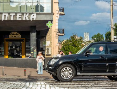Kyiv, Ukrayna - 27 Mayıs 2023: Parke sokağına park edilmiş siyah SUV, yoğun bir kentsel olay yerindeki bir eczane binasının yanındaki kaldırımda duran kadın