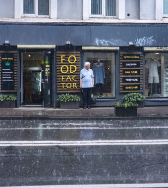 Kyiv, Ukrayna - 27 Mayıs 2023: Bir gıda dükkanı ve tekstil mağazasının yanındaki tentenin altında duran yaşlı adam, ıslak sokağa yağan şiddetli yağmurdan kaçınıyor.