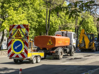 Yol onarım ekibinin ağır ekipmanlarla çalıştığı inşaat sahasında su tankı, kepçeli bir kamyon ve devam eden bakım için trafik işaretleri var.
