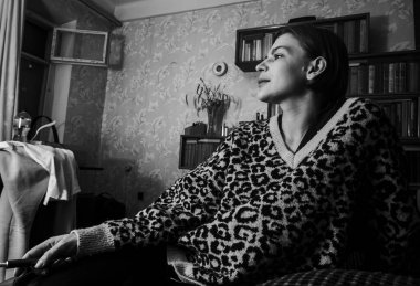 Kaliteli iç çamaşırlı genç bir kadın, leopar desenli bir kazakla pencerenin kenarında oturur, düşünceli, rahat ve düşünceli bir şekilde tek renkli bir ev sahnesine bakar.