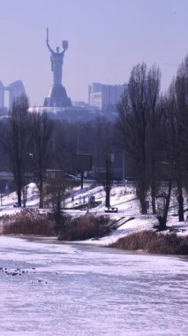 Donmuş bir su kütlesi ve karla kaplı bir parkın üzerindeki tepenin üzerinde duran ve Doğu Avrupa 'da soğuk bir kış günü arka planda görünen şehir manzarası ile birlikte anıtsal Ukrayna Ana heykeli.